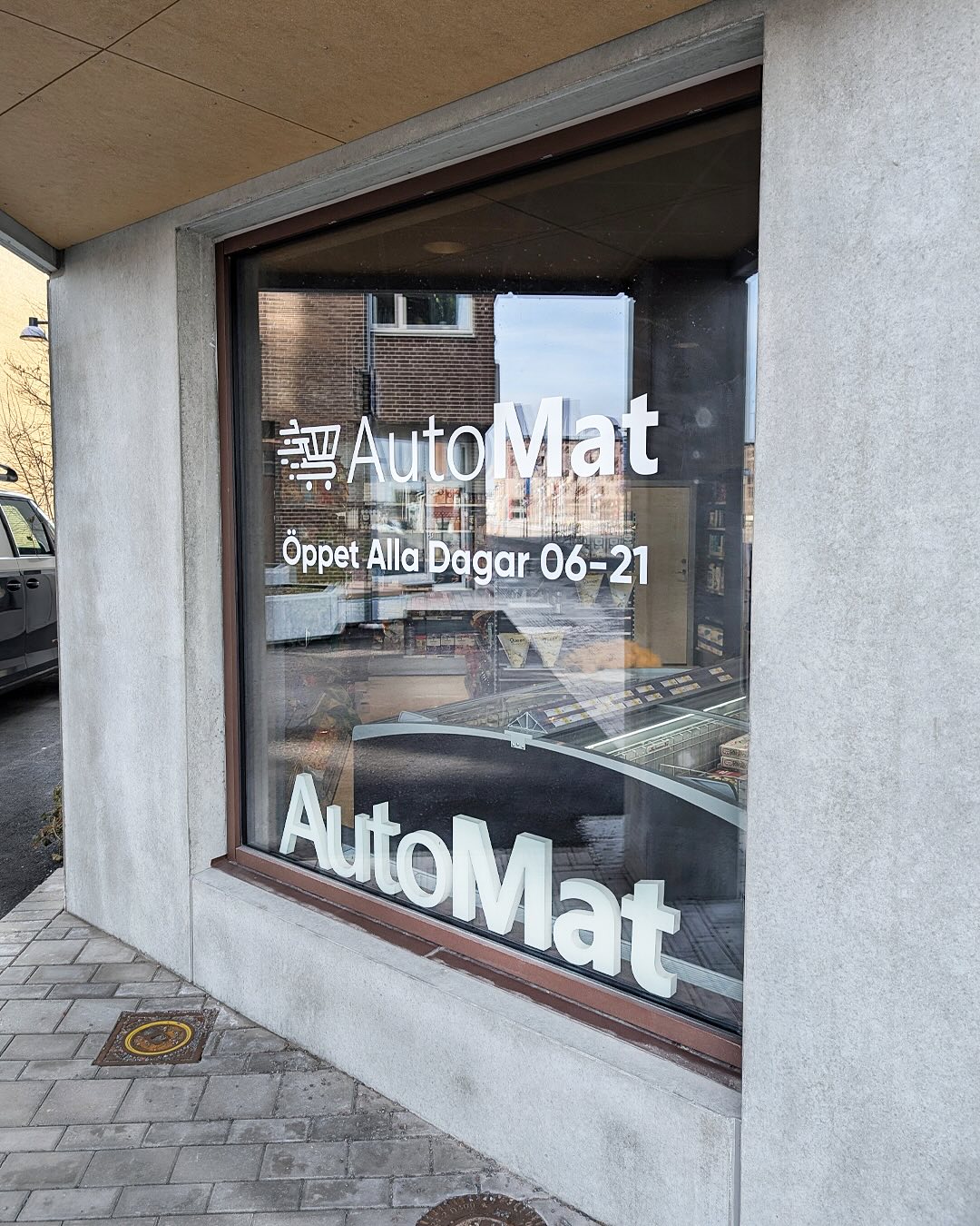 Skyltfönster med vit skuren dekor: kundvagnslogotyp, texten AutoMat och Öppet Alla Dagar 06-21, bakom glaset står fristående AutoMat-bokstäver