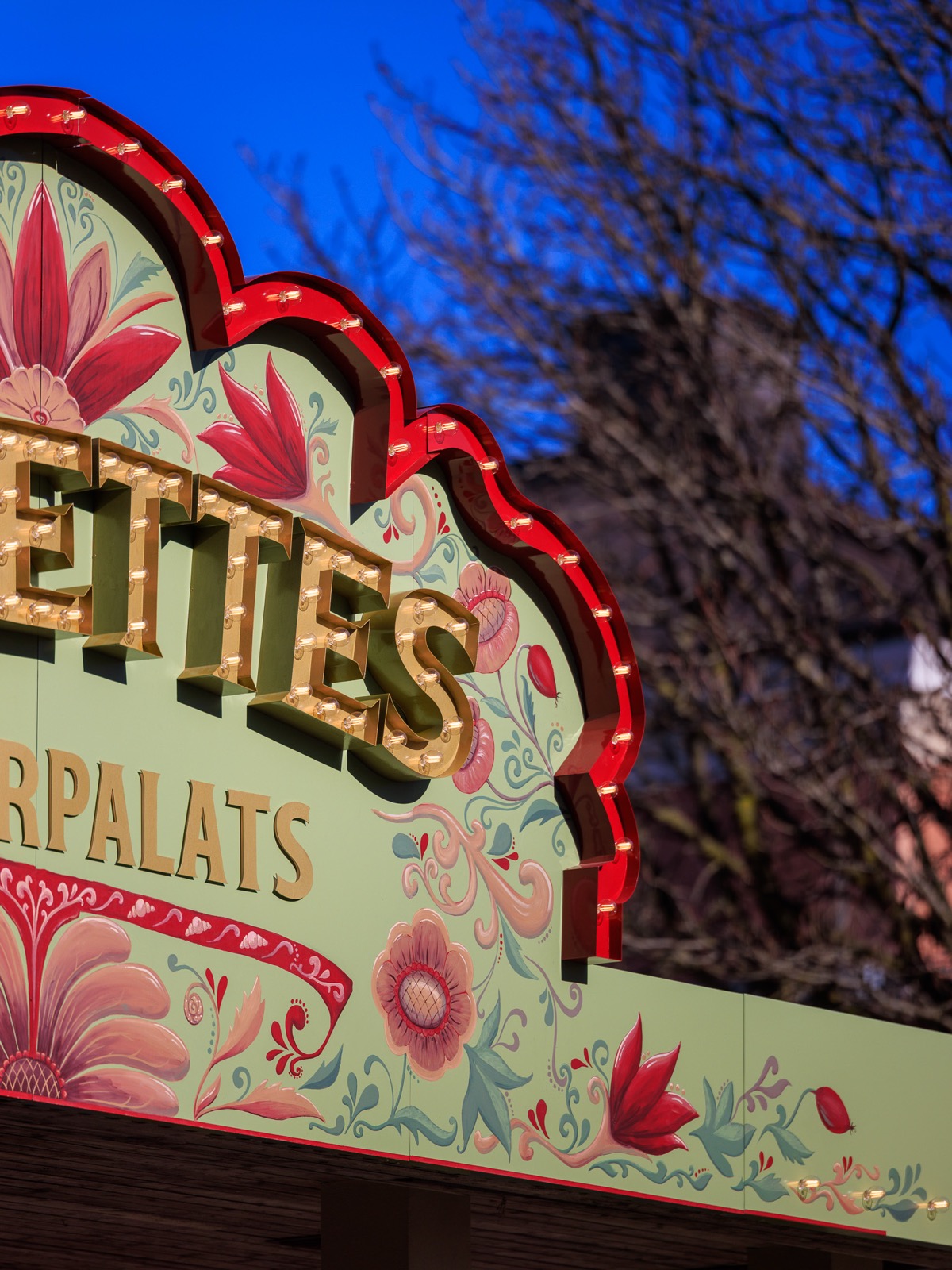Närbild på Babettes Kulturpalats entréskylt med handmålad blomsterdekor, guldbokstäver och ljusslingor