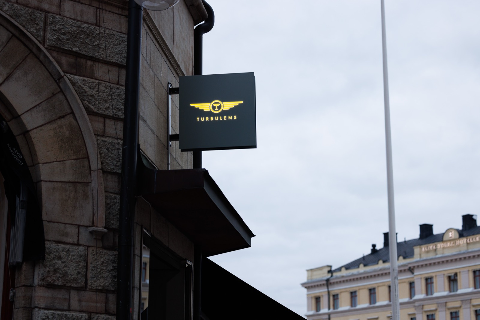 Svart pendelskylt där Turbulens logotyp lyser genomlyst i gult ur den mörka fronten