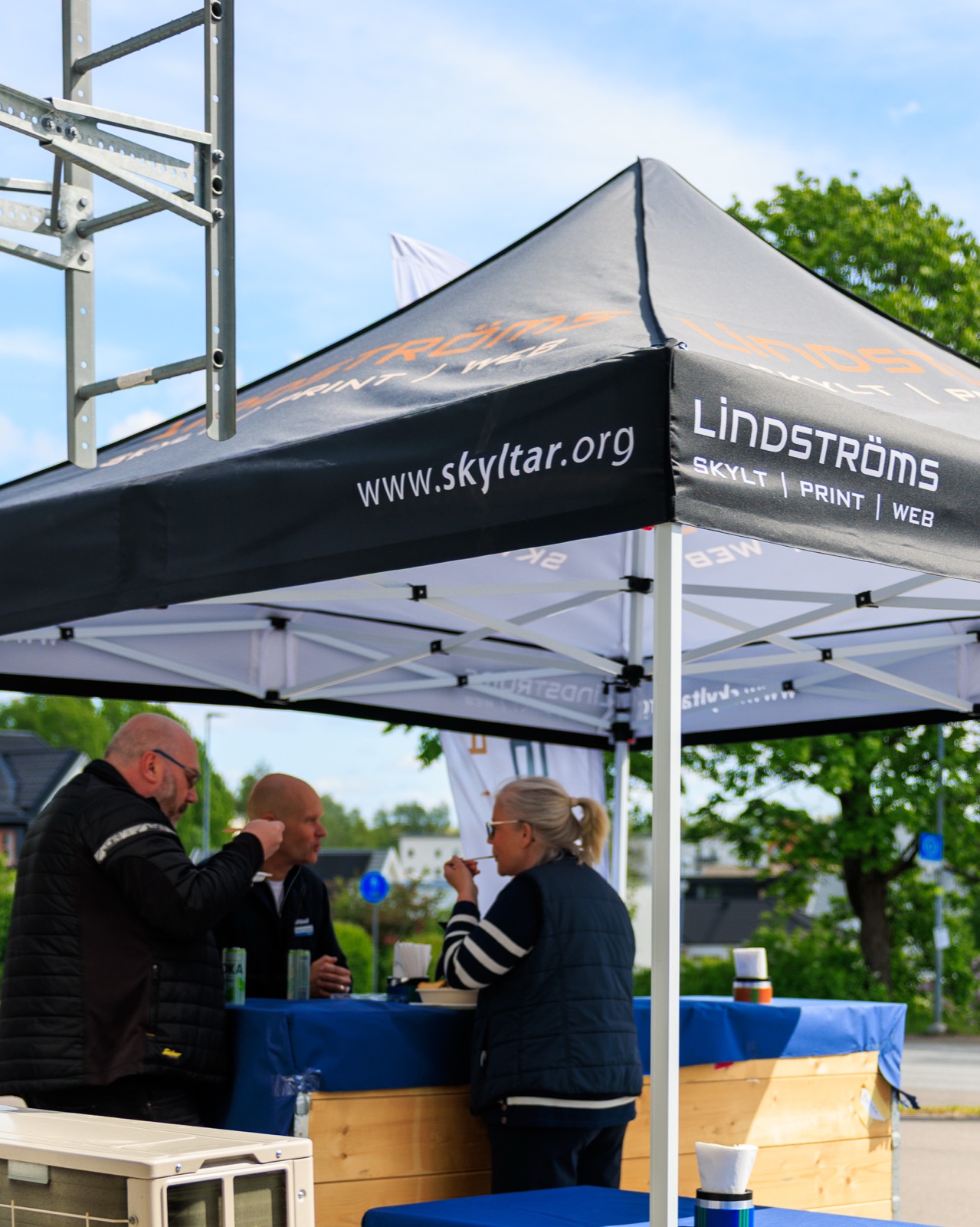 Lindströms svarta eventtält med logotyp och skyltar.org-tryck, tre personer under taket