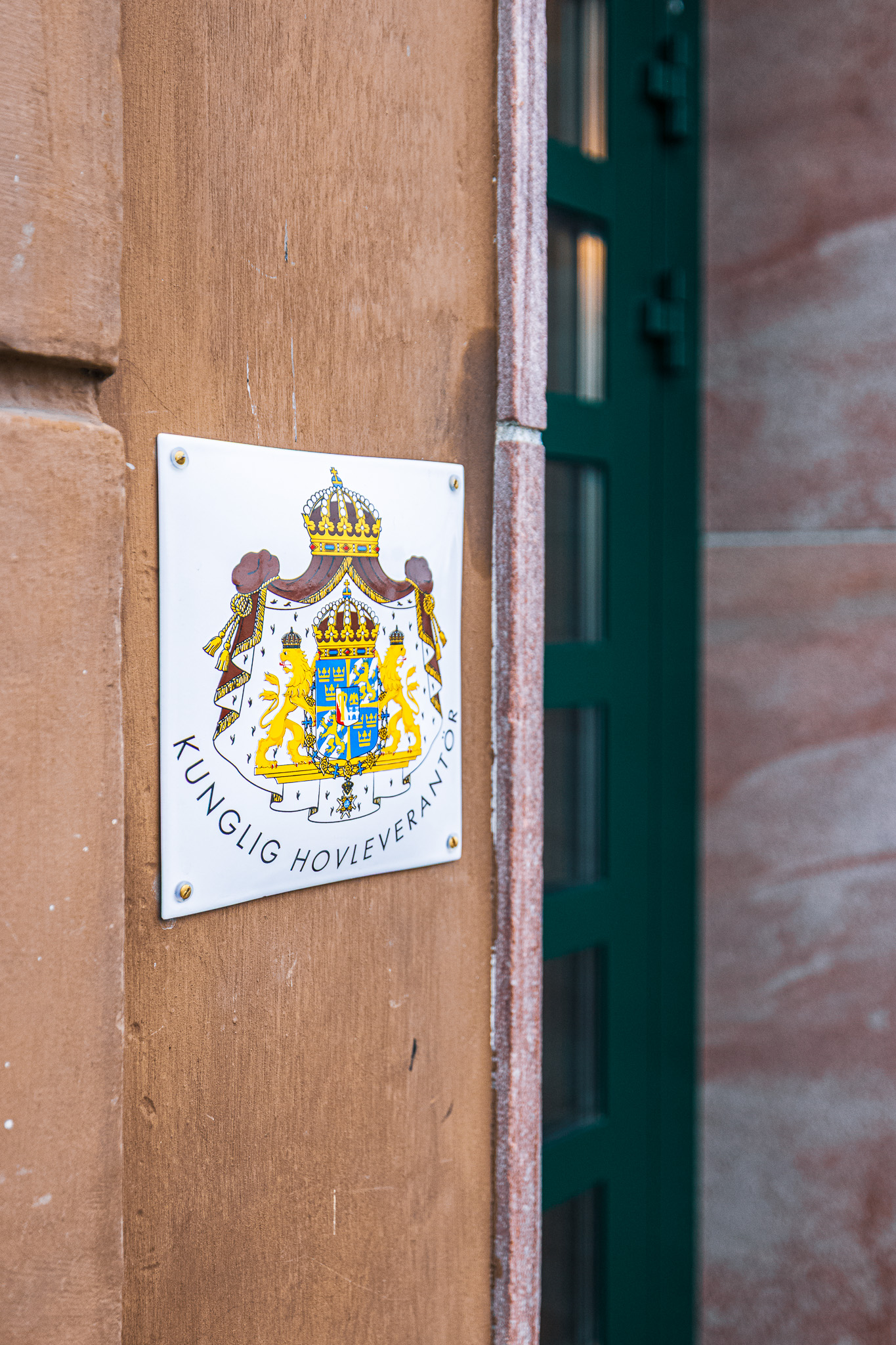 Vit emaljskylt med stora riksvapnet och texten Kunglig hovleverantör, skruvad på putsfasad intill en grön dörr
