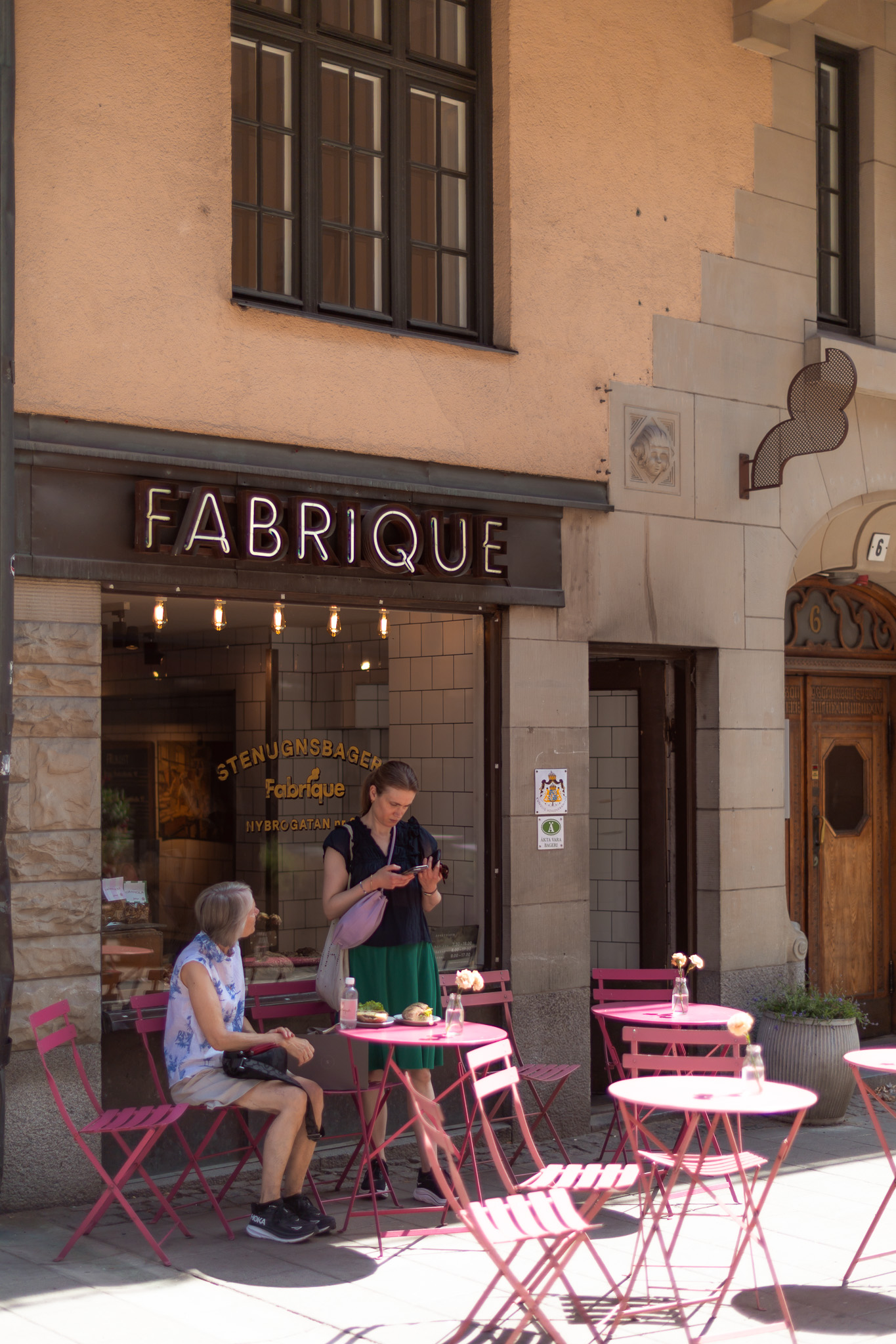 Fabriques butik på Nybrogatan en sommardag med neonskylt ovanför skyltfönstret, hovleverantörsskylten på fasaden och rosa cafébord på trottoaren