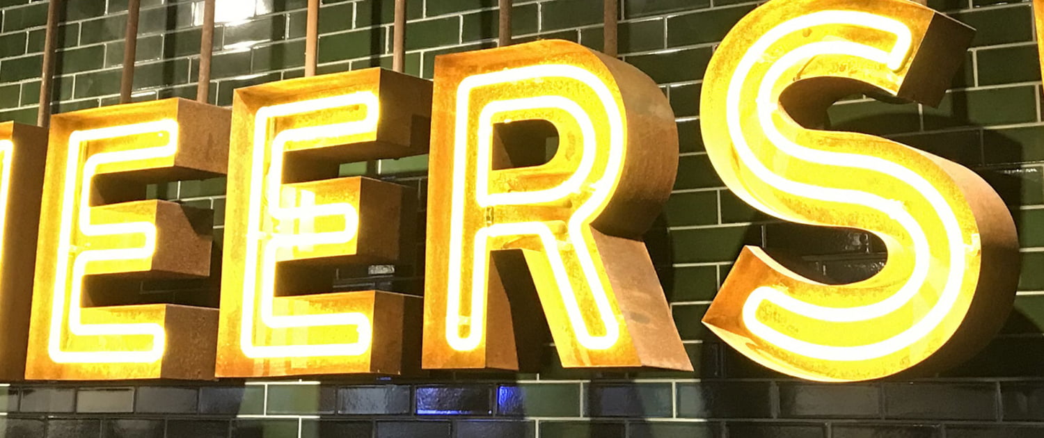 Upplyst neonskylt med texten Cheers mot grön tegelvägg