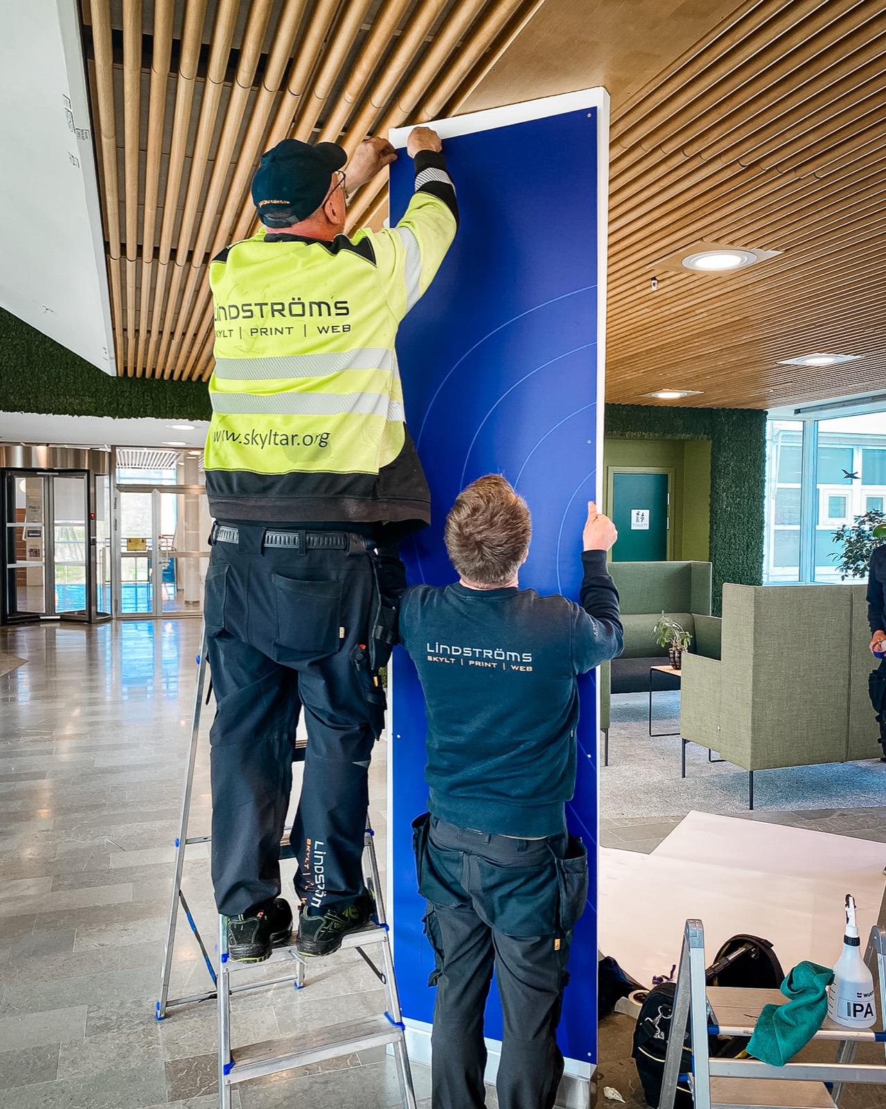Två montörer i Lindströms-kläder monterar en blå skyltpanel i en entréhall