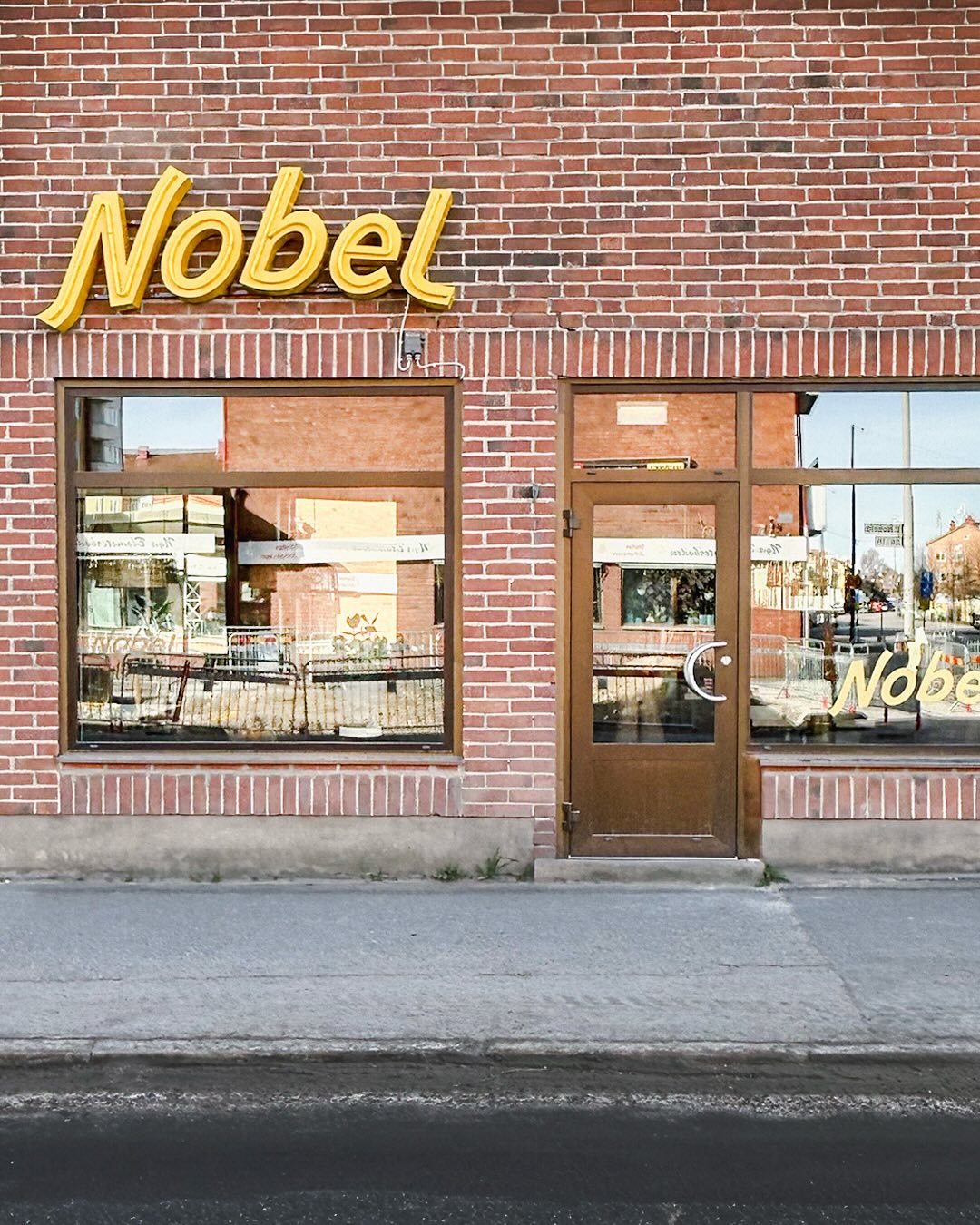 Gula bokstäver i skrivstil som bildar ordet Nobel på en tegelfasad, under dem skyltfönster med Nobel i guldtext på glaset