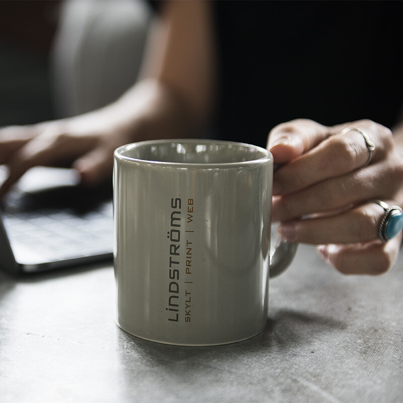 Grå keramikmugg med Lindströms logotyp i handen vid en laptop