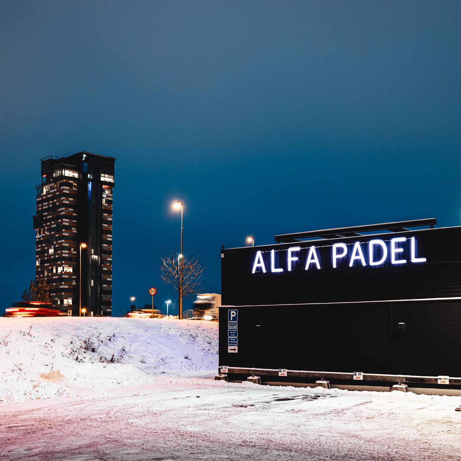 Belysta profilbokstäver med texten Alfa Padel på mörk fasad en vinterkväll