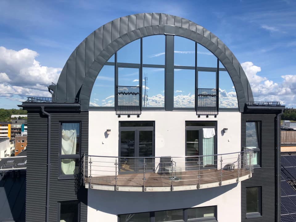 Bågformat glasparti högst upp på en byggnad, där de tonade rutorna speglar blå himmel och moln