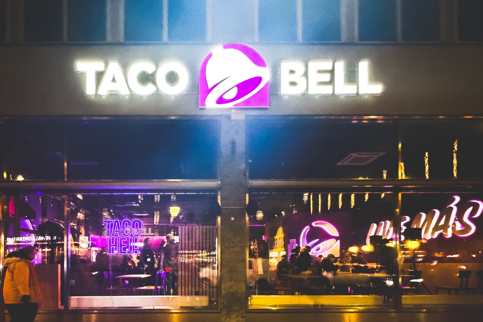 Taco Bells fasad i kvällsljus med belysta profilbokstäver, logotypskylt och neon i skyltfönstren