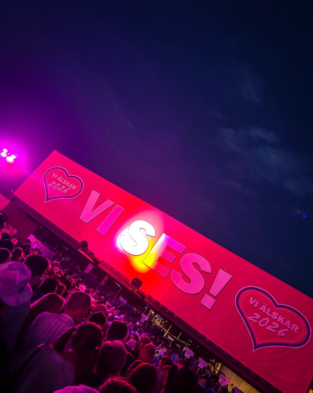 Stor rosa vepa med texten VI SES! upplyst i kvällsmörkret ovanför festivalpubliken